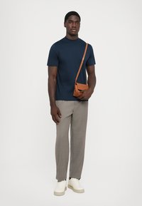 Tricou bleu marin cu mânecă scurtă, pantaloni gri în carouri și adidași albi; geantă de umăr maro cu curea. Fundal neutru.