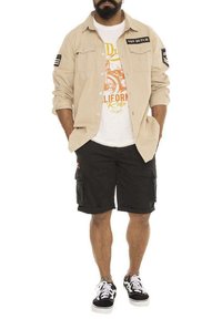 Chemise beige à boutons avec des écussons, t-shirt blanc avec impression orange, shorts cargo noirs, et baskets noires avec semelles blanches.