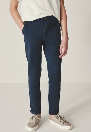 Person trägt schlanke marineblaue Hose mit hochgekrempelten Bündchen, ein weißes Hemd, das hinein gesteckt ist, und hellbeige Sneakers mit gebundenen Schnürsenkeln.