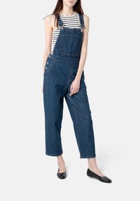 Pantalones overoles de mezclilla con un corte holgado, que presentan tirantes ajustables, un bolsillo frontal, detalles de botones en los laterales y piernas cortas. Combinados con una blusa a rayas.