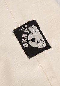 Beżowy materiał z kwadratową czarną metką, na której znajduje się biała, rysunkowa dłoń wykonująca gest "OK" oraz napis "W PORZĄDKU" w zabawnym kroju pisma.
