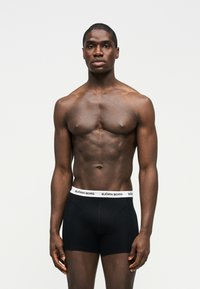 Μυώδης άνδρας που στέκεται μπροστά σε απλό φόντο, φοράει μαύρα μπόξερ με λευκή μέση και την επιγραφή "BJÖRN BORG", με τα χέρια χαλαρά.