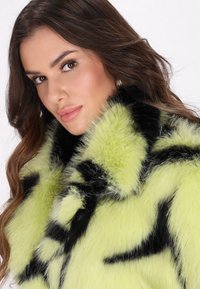 Cappotto di pelliccia sintetica verde con motivi neri, caratterizzato da un colletto alto e una texture morbida. Primo piano dei dettagli del tessuto e del contrasto di colori.