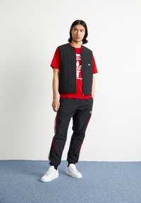 Homem com uma t-shirt vermelha da Adidas, colete acolchoado preto, calças de tracksuit pretas da Adidas com riscas vermelhas e ténis brancos, de pé sobre um tapete azul.