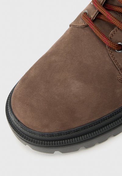 Botte de randonnée en cuir suédé marron avec semelle en caoutchouc noir et lacets marron-rouge sur fond blanc, montrant un gros plan de la zone des orteils.
