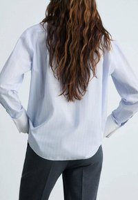 Camicia azzurra chiara a maniche lunghe con sottili righe verticali, vestibilità rilassata, polsini con bottoni e orlo curvo, abbinata a pantaloni grigio scuro.