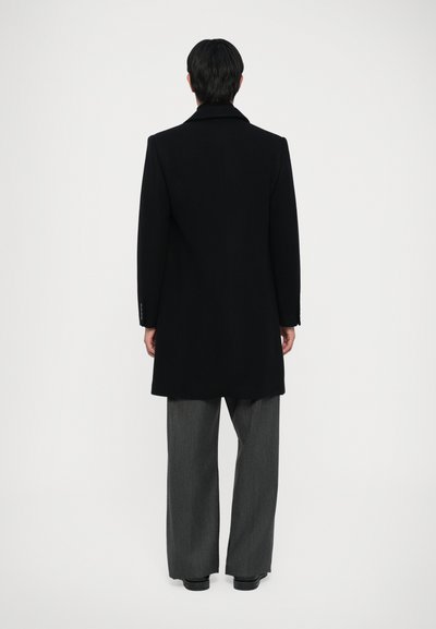 Cappotto lungo nero con colletto a punte, vestibilità sartoriale e spacco posteriore, abbinato a pantaloni grigi a gamba larga. Texture liscia e design minimal.