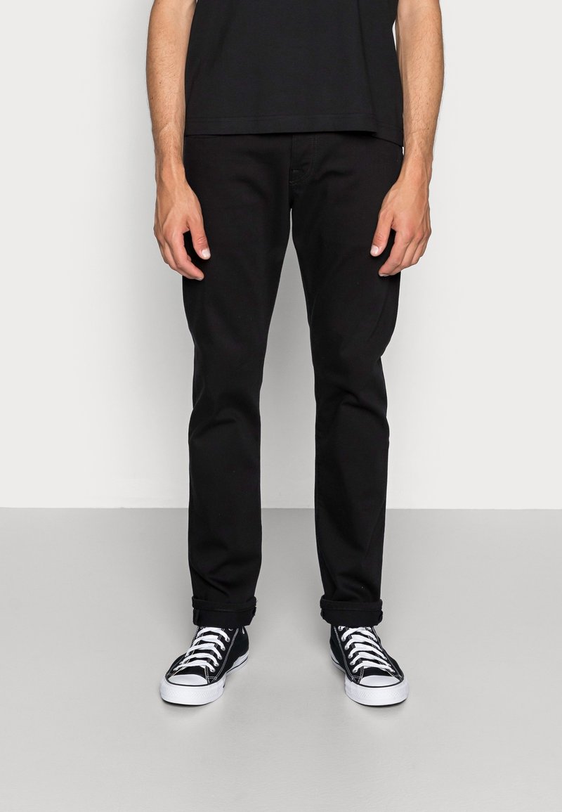Pantalones de mezclilla negros con corte ajustado, que presentan un diseño clásico de cinco bolsillos, bajos enrollados, combinados con zapatillas de lona bajas en blanco y negro.