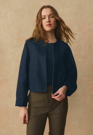 Junge Frau mit langen Haaren, die eine marineblaue Jacke mit Reißverschluss und braune Hose trägt, vor einem beigen Hintergrund stehend.