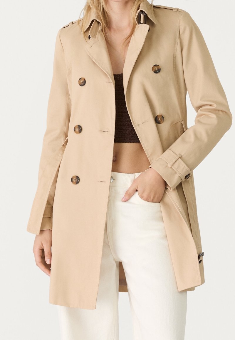 Beige trenchcoat met dubbele rij knopen, schouderepauletten en zijzakken. Gemaakt van lichtgewicht materiaal; valt boven de knie.