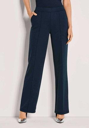 Vrouw in marineblauwe op maat gemaakte rechte broek en metallic zilveren puntige hakken, met één hand in de zak.