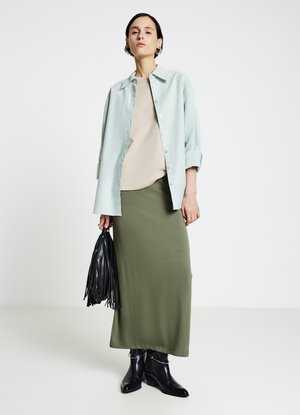 Chemise boutonnée bleu clair, haut beige, jupe vert olive, bottines noires, tenant un sac à franges noir. Design simple, textures lisses.