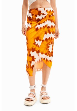 Falda de tie-dye con patrones en naranja y marrón, tela suave, dobladillo asimétrico y detalle de cintura anudada. Llevada con sandalias de plataforma blancas.