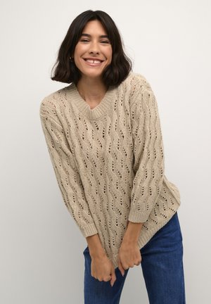 Maglione beige lavorato a maglia con scollo a V, caratterizzato da un motivo a trafori e maniche lunghe. Abbinato a jeans in denim blu.