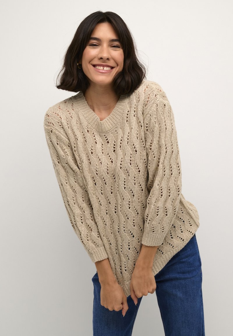 Maglione beige lavorato a maglia con scollo a V, caratterizzato da un motivo a trafori e maniche lunghe. Abbinato a jeans in denim blu.