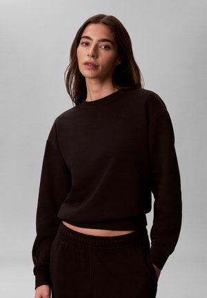 Femme aux longs cheveux bruns portant un sweat court noir et un pantalon noir assorti, debout devant un fond uni gris clair.