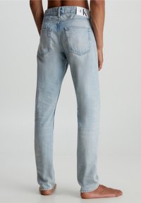 Světle modré džíny s rovný fit, pěti kapsami a bílou visačkou na pasu s logem značky. Poškozená textura poblíž dolního okraje.