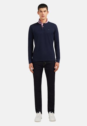 Homme debout portant un polo à manches longues navy avec un col rose, un jean foncé et des baskets blanches, sur un fond blanc.