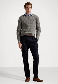 Suéter cinzento de malha com cabo, camisa azul com colarinho, calças azul-marinho com riscas finas, sapatos de couro castanhos, pose em pé sobre um fundo neutro.