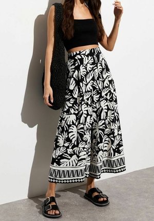 Femme portant un crop top noir, un pantalon large à motif feuilles noir et blanc, des sandales noires et un grand sac tote noir en tricot.