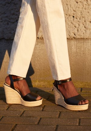 Czarne skórzane sandały na koturnie z otwartym palcem, pleciona platforma z juty, regulowany pasek w kostce, noszone z jasnobeżowymi spodniami na fakturowanej powierzchni.