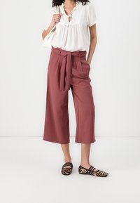 Bordeaux wijde broek met een strikceintuur, gecombineerd met een witte textuur blouse en luipaardprint flats. De achtergrond is eenvoudig wit.