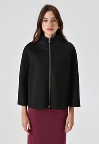 Chaqueta negra con capa, cuello alto y cremallera frontal, con un tirador de cremallera dorado, fabricada en tejido suave y con mangas fluidas, llevada sobre una falda burdeos.