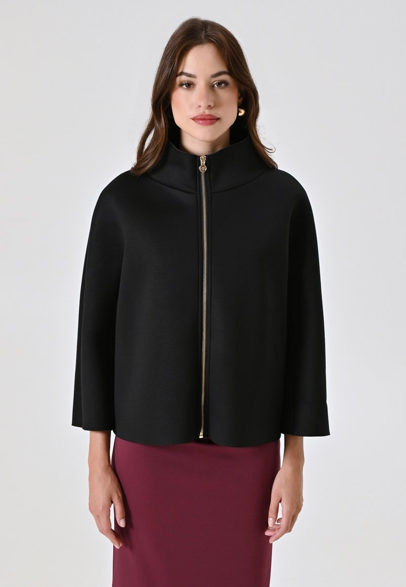 Chaqueta negra con capa, cuello alto y cremallera frontal, con un tirador de cremallera dorado, fabricada en tejido suave y con mangas fluidas, llevada sobre una falda burdeos.
