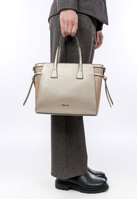 Borsa a mano in oro metallico con finitura testurizzata, doppi manici corti, chiusura con zip e dettagli beige, tenuta da una persona in pantaloni grigi su misura.