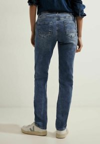 Jeans de mezclilla azul oscuro con corte recto, cinco bolsillos y un leve desgaste en las piernas, combinados con zapatillas blancas.