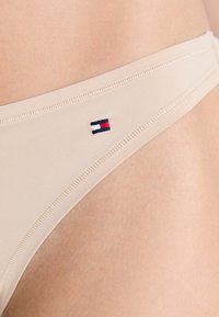 Bežinės bikini kelnaitės su mažąja Tommy Hilfiger vėliavėlės logo šone, dėvimos ant žmogaus klubo ir viršutinės šlaunies srities.