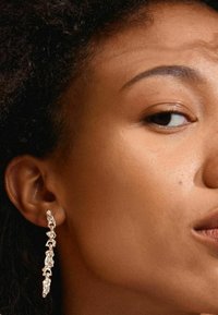 Pendientes de plata con un diseño abstracto y alargado que presenta varias facetas texturizadas, colgando por debajo del lóbulo de la oreja sobre un cabello oscuro.