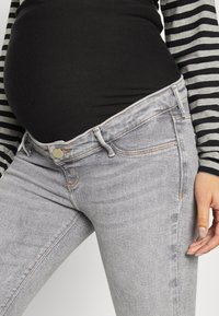 Pantalones vaqueros de maternidad en denim gris claro con un diseño clásico de cinco bolsillos. Cuentan con cierre de botón y un ligero desvanecimiento a lo largo de los muslos.