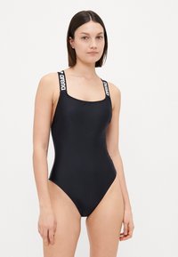 Maillot de bain une pièce noir avec encolure carrée, larges bretelles portant le logo "arena" et une texture lisse et élégante.
