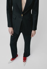 Person wearing a black blazer buttoned once over bare chest, matching black pants with front slits, red socks, and white pointed shoes.