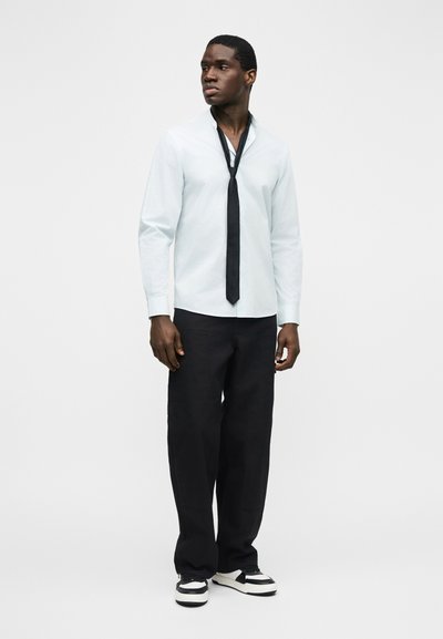 Άνδρας στέκεται φορώντας λευκό πουκάμισο χωρίς να είναι μανταρισμένο, φαρδιά μαύρη παντελόνια, μαύρη γραβάτα χαλαρά δεμένη και ασπρόμαυρα αθλητικά παπούτσια, κοιτάζοντας προς τα αριστερά.