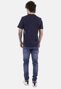 Mann mit lockigem Haar, tätowiertem rechtem Arm, trägt ein marineblaues T-Shirt, blaue Jeans und weiße Sneakers, steht mit dem Rücken zur Kamera vor einem weißen Hintergrund.