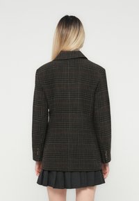 Persoon met lang blond haar draagt een donker geruite blazer en een geplooide donkere rok, staande voor een eenvoudige witte achtergrond.