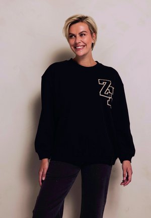 Black sweatshirt featuring a prominent gold "Z" logo. Oversized fit with wide sleeves and a rounded neckline. Soft fabric with a casual design.