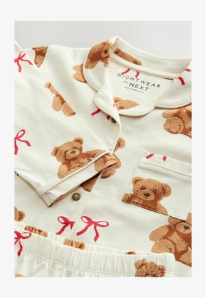 Pijamale din bumbac cu un fundal crem, decorate cu ursuleți de pluș și fundițe roșii. Include o buzunar și închidere cu nasturi pe partea de sus.