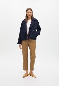 Trench-coat bleu marine croisé, pantalon beige et ballerines dorées. Le manteau a de gros boutons et un col surdimensionné ; le pantalon est ajusté.