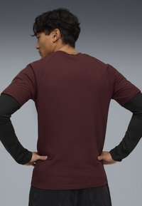 Camisa corta de manga marón con una textura suave. Presenta una camiseta de manga larga negra visible en las mangas y un diseño de ajuste relajado.