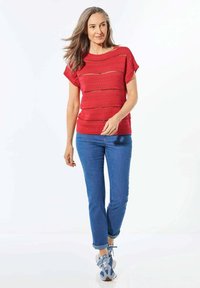 Rode gebreide top met korte mouwen, voorzien van horizontale patronen en uitsnijdingen, gecombineerd met blauwe opgerolde jeans en patroon sneakers.