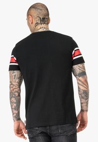 T-shirt nera di cotone a maniche corte con strisce rosse e bianche. I tatuaggi visibili su braccia e collo completano il look.