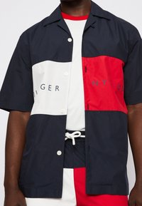 Camicia a maniche corte in tessuto leggero, con pannelli blu navy, bianco e rosso e grande stampa del marchio. Dettagli distintivi su bottoni e colletto.