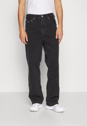 Homme portant un jean noir ample et des baskets blanches, debout devant un fond clair uni, mains détendues sur les côtés.