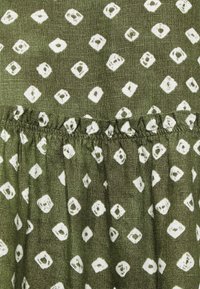 Gröntyg med en texturerad yta som har ett upprepande mönster av vita diamanter och rutor. Inkluderar en rynkad detalj i kanten längst ner.