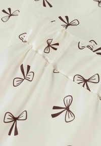 Witte stof met donkerbruine strikpatronen en een gathered elastische naaddetail in het midden, met een close-upweergave van de textiel.