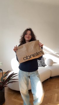 Rjava papirnata vrečka z napisom "zalando" v debelih, črnih črkah, ki jo drži oseba. Osvetlitev in strukturiran bel kavč v ozadju.