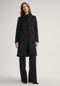 Zwarte wollen mixjas met een hoge kraag, zes knopen aan de voorkant, zijzakken en een knielange silhouet. Soepele textuur, tailormade ontwerp.